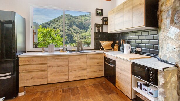 Eine moderne Küche mit Holzschränken, einem schwarzen Kühlschrank, einer Wand mit Steinverkleidung und Blick auf eine Berglandschaft.