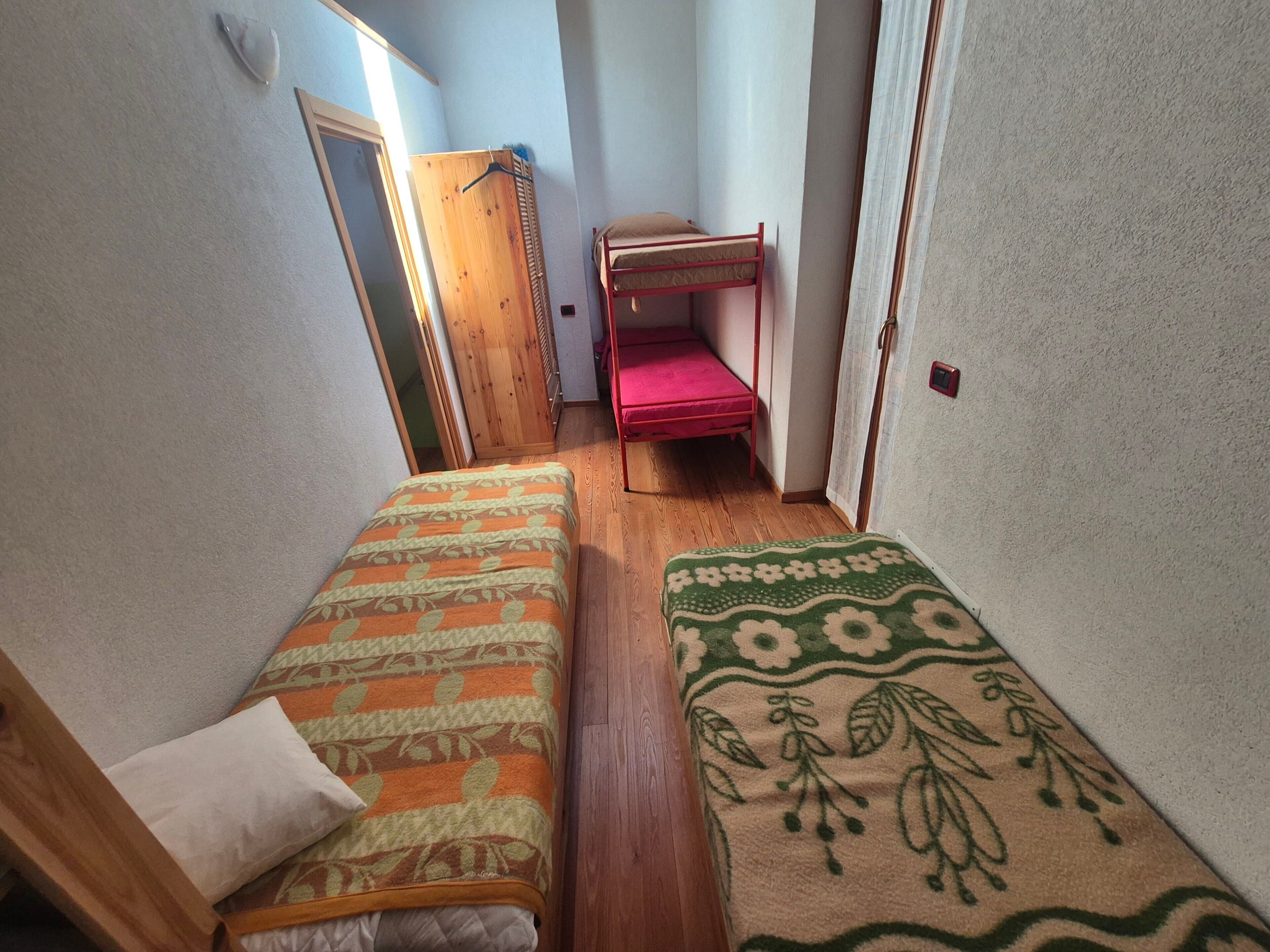 A narrow room with two beds, a wooden wardrobe, and a red bench.