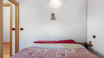Een slaapkamer met een bed, een aan de muur bevestigd religieus beeldje, een deur en een nachtkastje met een lamp.
