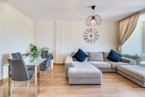 A modern living room with a glass dining table, grey sofas, and a wall clock.