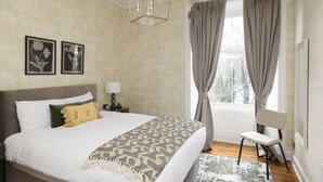 Un dormitorio con cama, una silla, una mesita de noche, una lámpara y una ventana con cortinas.