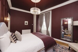 A bedroom with a large bed, a chandelier, a mirror, and a view of a bathroom.