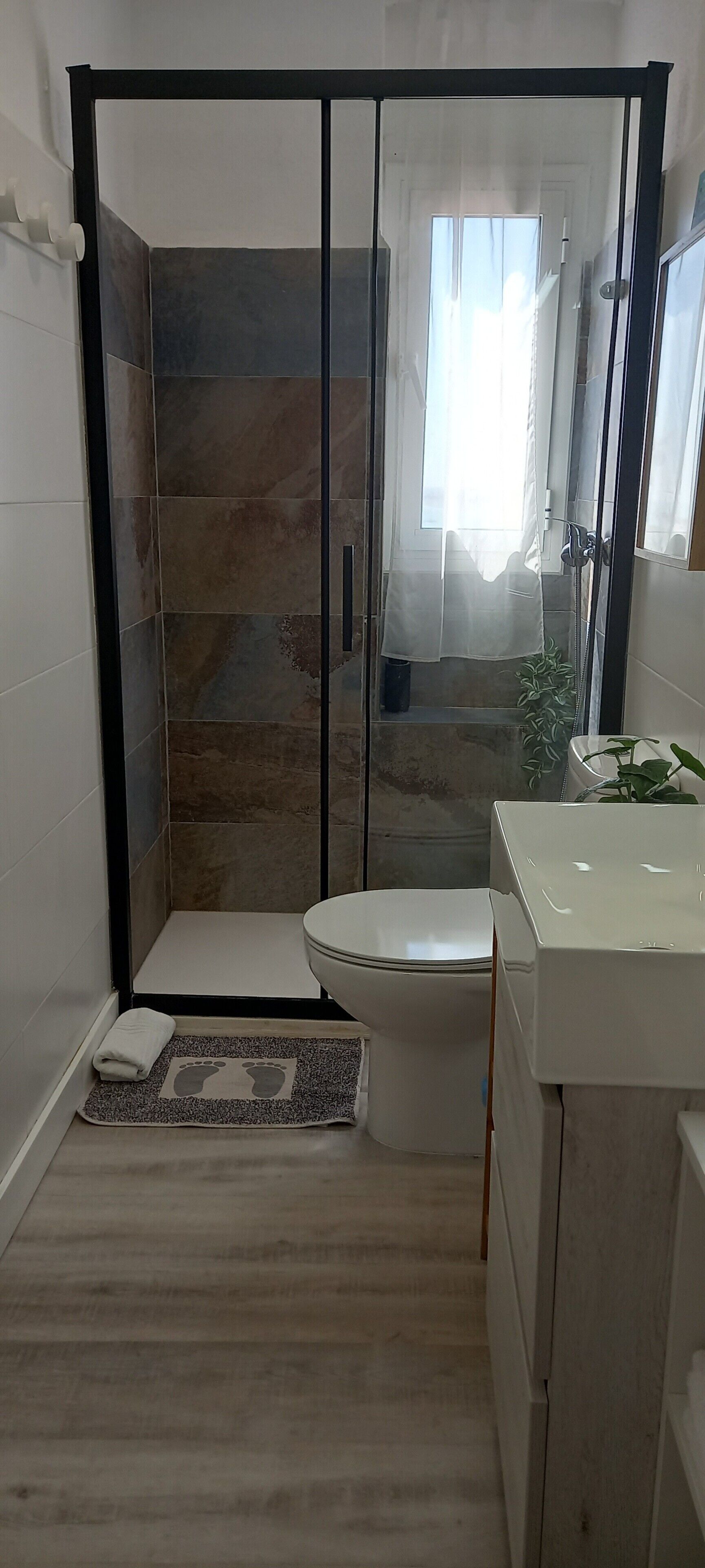 A bathroom with a shower enclosure, a toilet, a sink, and a small rug with a foot pattern.