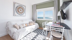Een moderne kamer met een witte bank, een eettafel met stoelen, een aan de muur bevestigde televisie en uitzicht op zee.