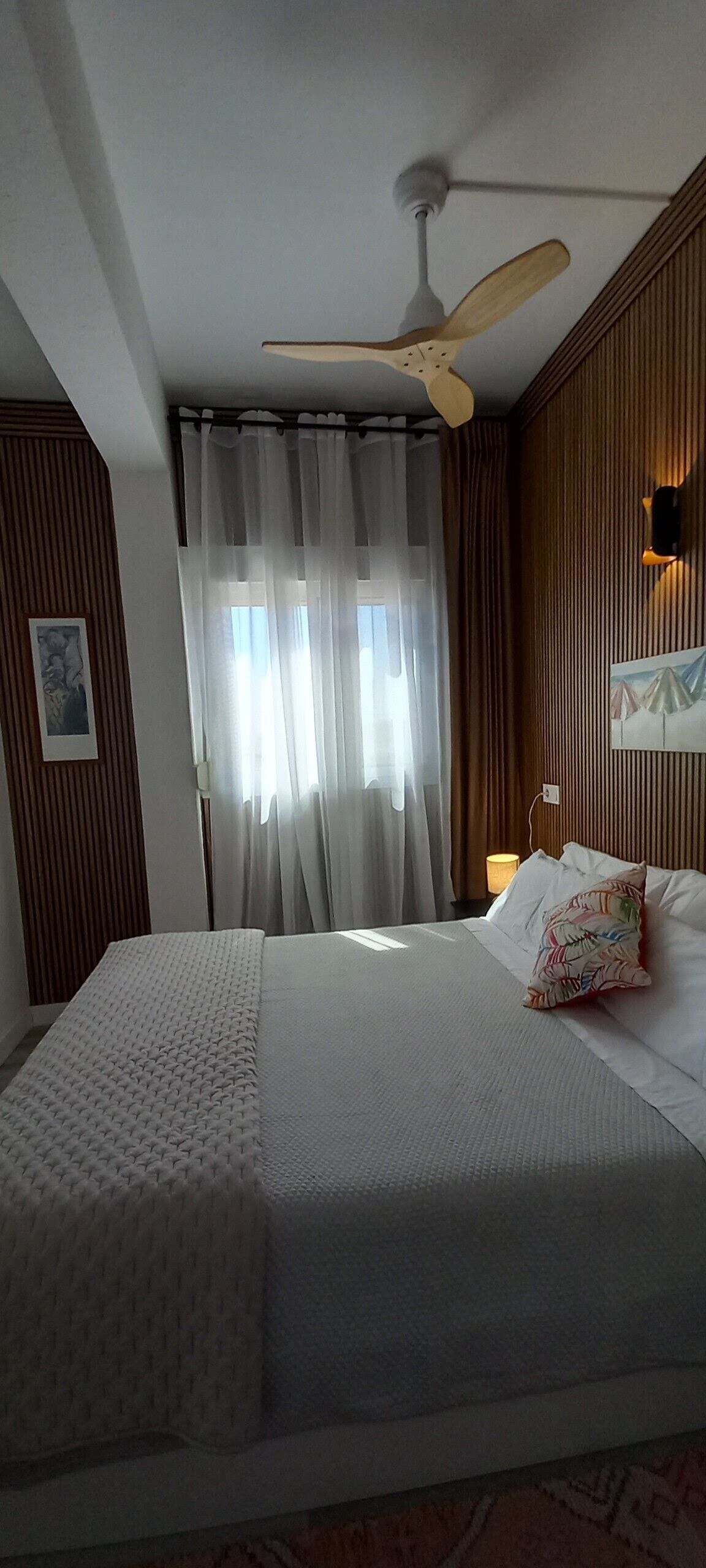 A hotel room with a bed, a ceiling fan, a window with curtains, a wall-mounted light, and a framed picture.