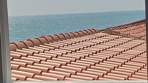 Uitzicht door een raam op een met tegels bedekt dak, mogelijk een balkon of terras, met een heldere lucht en een verre horizon.