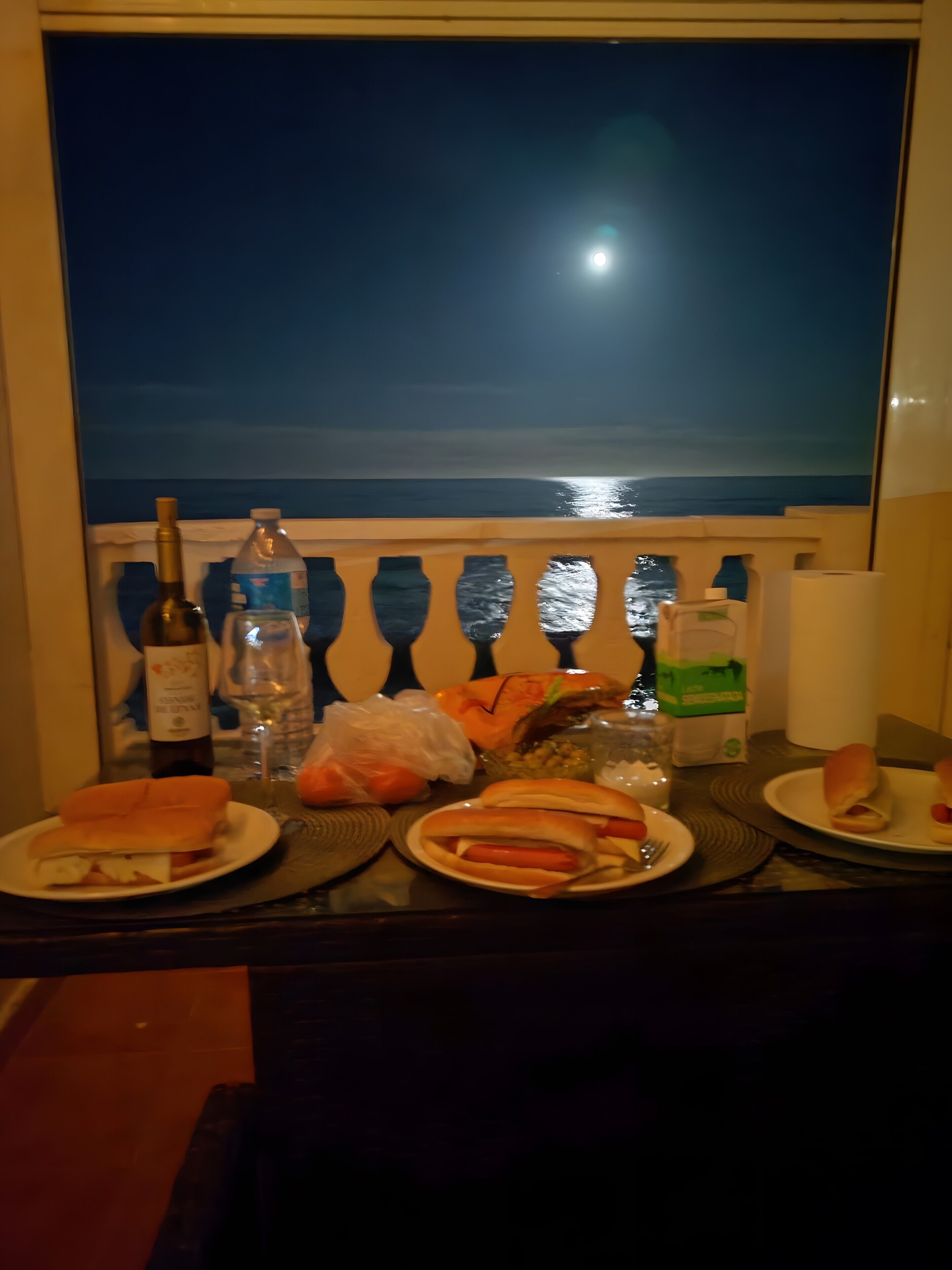 Een tafel met eten en drinken, een fles wijn, en uitzicht op de oceaan 's nachts.