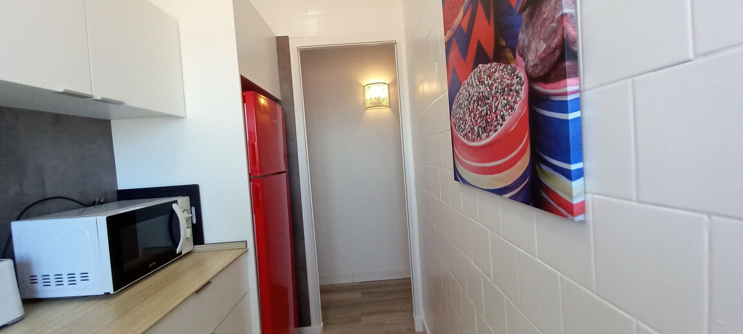 A narrow hallway with a microwave on a counter, a red refrigerator, and a wall-mounted light fixture.