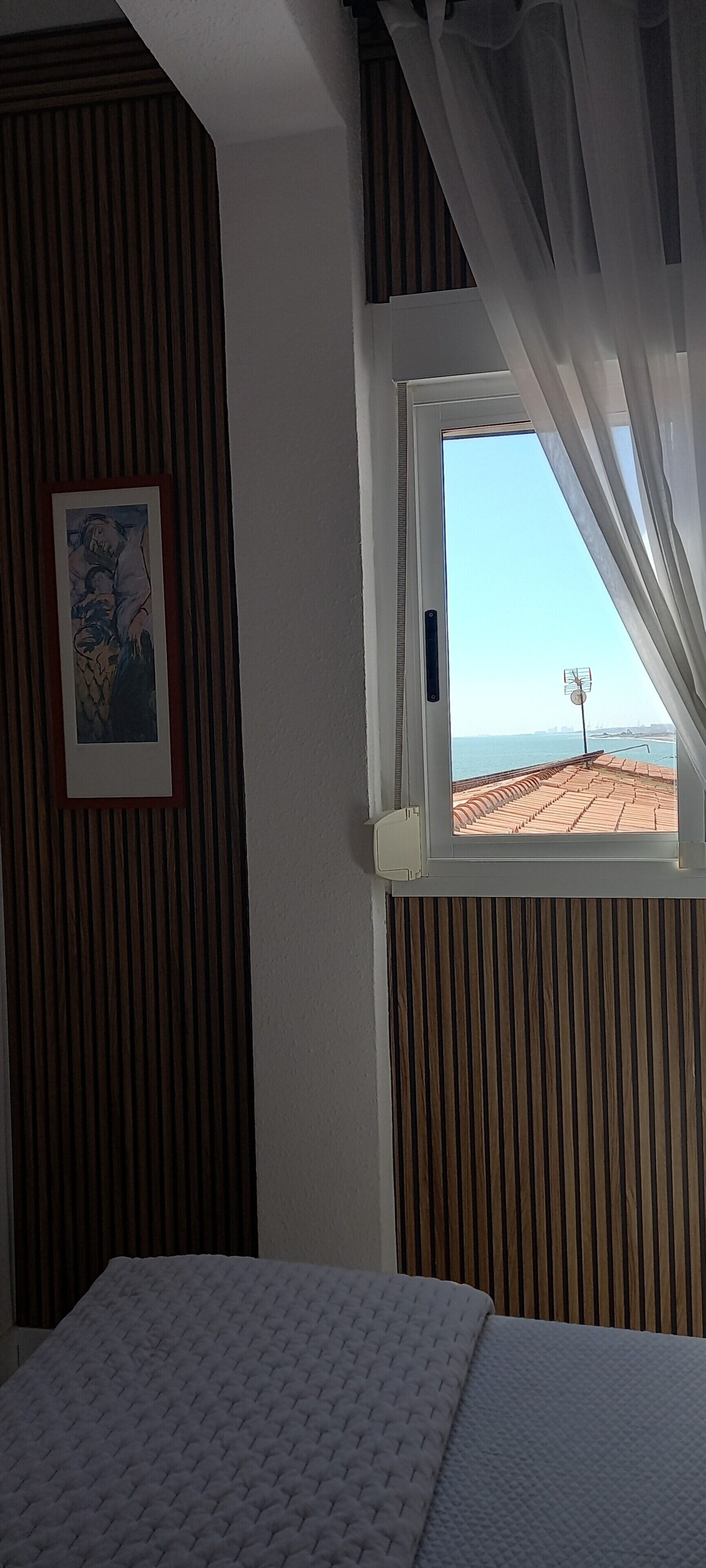 Una habitación con cama, ventana con cortinas, un cuadro en la pared y vista al mar.