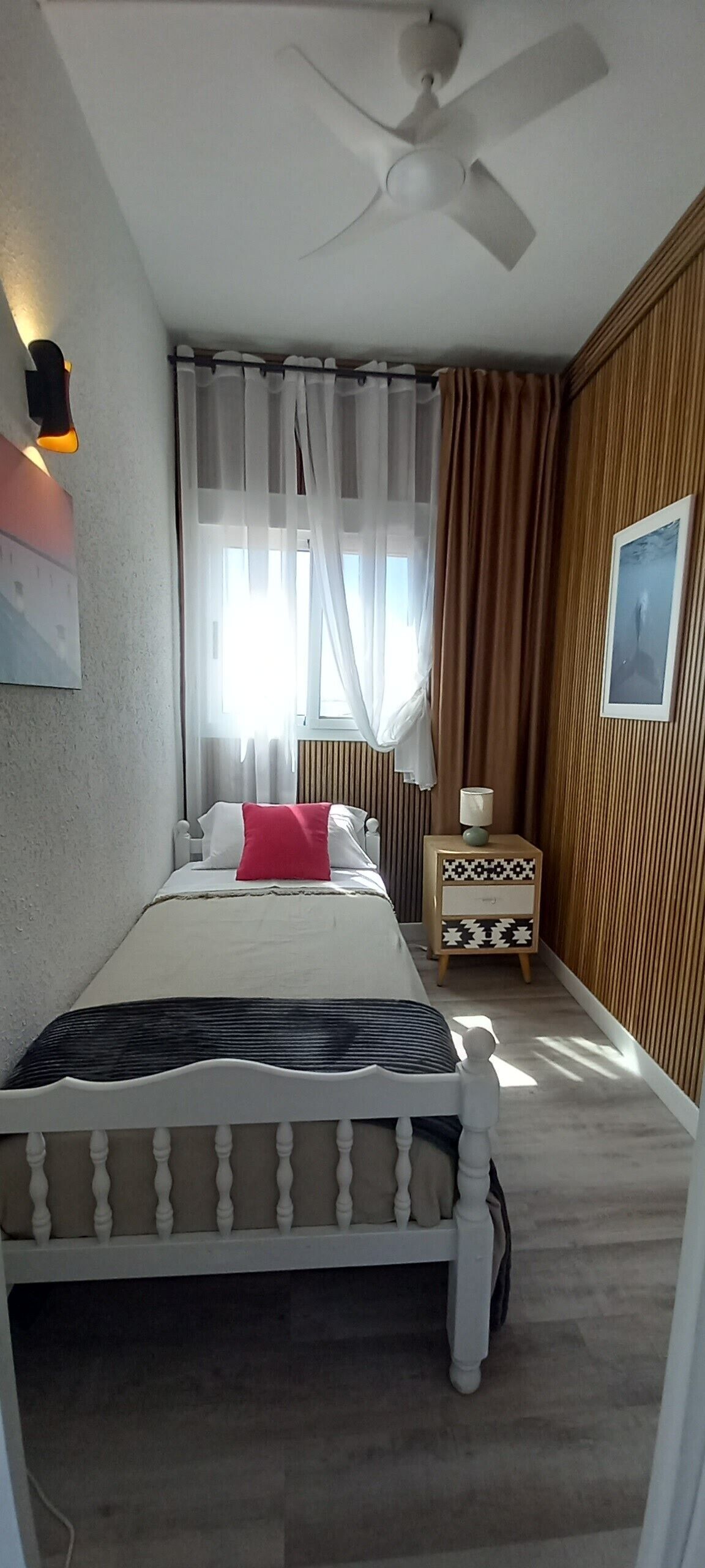 Quarto com cama, mesa de cabeceira, ventoinha no teto, janela com cortinas e um quadro na parede.