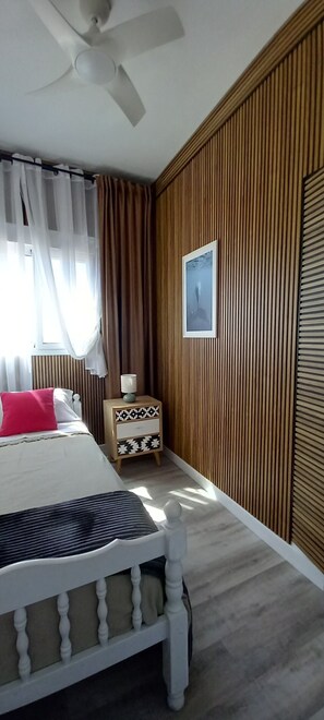 Quarto com cama, mesa de cabeceira, ventilador de teto, janela com cortinas e painel de parede de madeira.