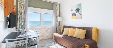 A room with a bed, a table with chairs, a TV, and a window with a view of the sea.