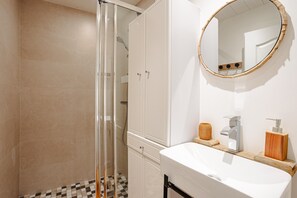 Ein Badezimmer mit einer Duschkabine aus Glas, einem weißen Waschbecken mit Seifenspender aus Holz und einem runden Spiegel an der Wand.