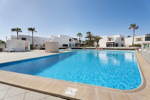 A swimming pool with a clear blue water, surrounded by white buildings, palm trees, and a stone wall. - Vacation Home 'Casa Relax Marine' with Shared Pool, Wi-Fi and Air Conditioning (Costa del Silencio Arona)