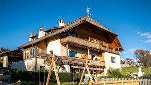 A two-story wooden house with a balcony, a swing set, and a stone wall in the foreground.