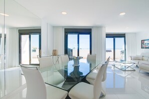 A modern living room with a glass dining table, white chairs, a sofa, and large windows with curtains.