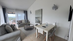 A modern living room with a sofa, a round coffee table, a dining table with chairs, a TV mounted on the wall, and a view through the window. - Apartment 'Appartamento Sea View' with Shared Pool, Private Terrace and Air Conditioning (Arzachena)