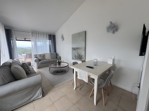 A modern living room with a sofa, a round coffee table, a dining table with chairs, a TV mounted on the wall, and a view through the window. - Apartment 'Appartamento Sea View' with Shared Pool, Private Terrace and Air Conditioning (Arzachena)
