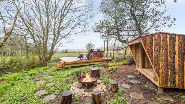 A rustic outdoor area with a wooden deck, two lounge chairs, a fire pit, and a wooden structure.