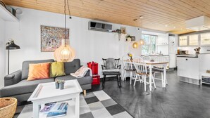 A modern living room with a grey sofa, a dining area with white chairs, and a kitchen with white cabinets.