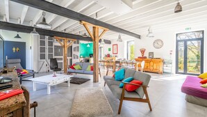 Un amplio salón con techo alto, vigas de madera, un sofá con cojines de colores, una mesa de centro y un comedor al fondo.