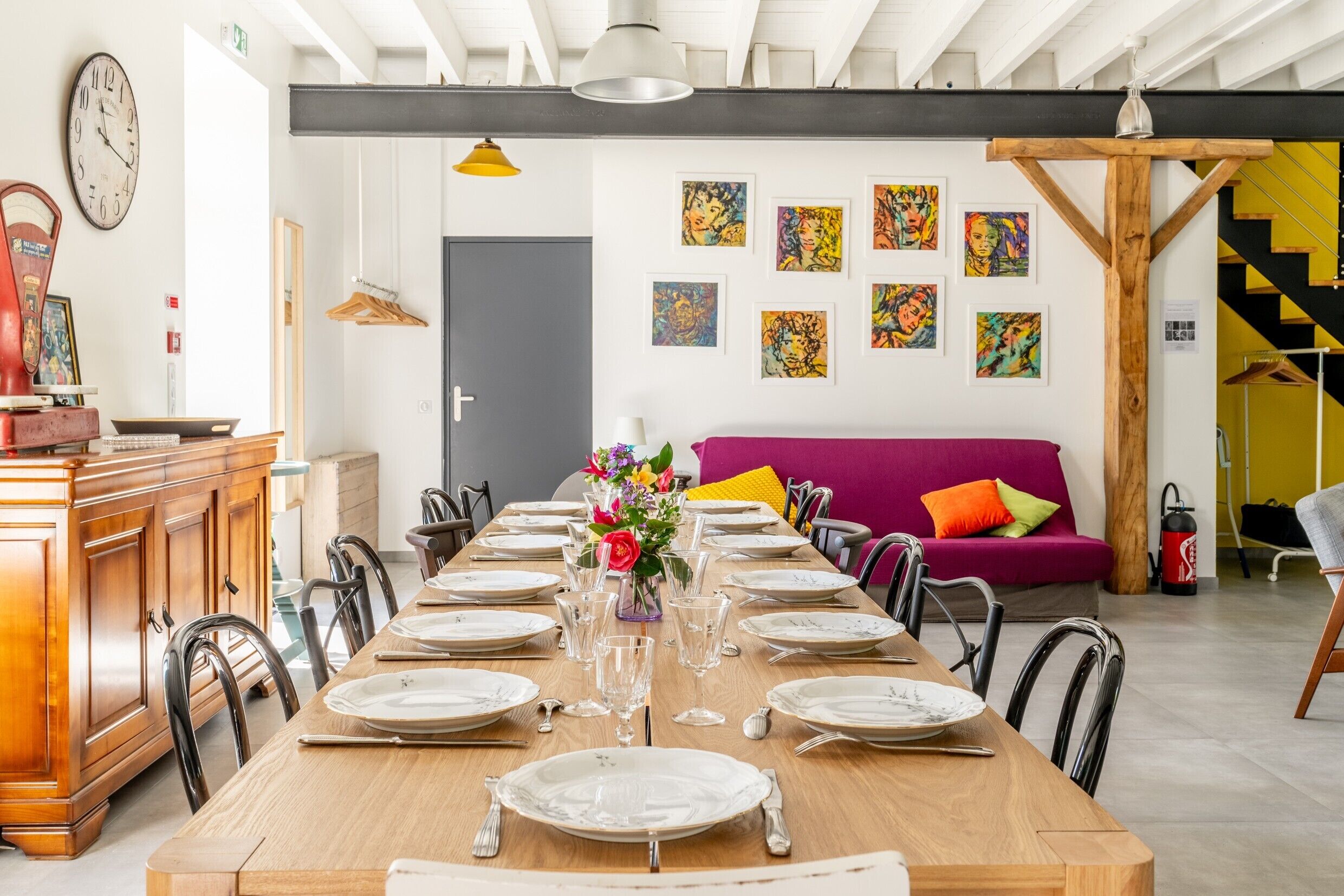 A dining area with a long wooden table set for a meal, a sofa with colorful cushions, and a staircase with a yellow railing.