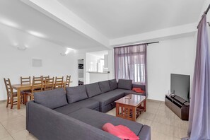 Un salon moderne comprenant un canapé d’angle, une table à manger avec des chaises, un téléviseur et une grande fenêtre avec des rideaux.