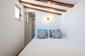 Quarto com cama, duas almofadas estampadas, mesa de cabeceira, luminária na parede e janela.