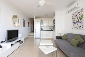 A modern living room with a flat-screen TV, a sofa with green cushions, a small white table, and a kitchen in the background.