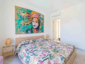 A bedroom with a large bed, a wall-mounted painting, a wicker chair, and a small table.