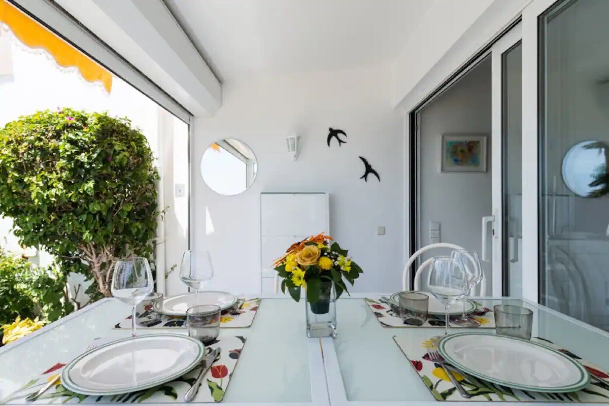 A dining area with a table set for two, a vase with yellow flowers, and a view of a tree outside.