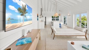 Una habitación amplia con un televisor grande que muestra una escena de playa, un banco de madera y vistas al océano a través de amplios ventanales.