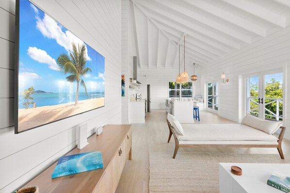 Una habitación amplia con un televisor grande que muestra una escena de playa, un banco de madera y vistas al océano a través de amplios ventanales.