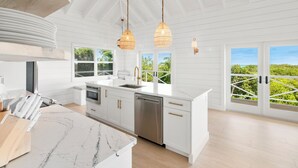 Cocina moderna con armarios blancos, encimera de mármol y lavavajillas. Amplias ventanas con vistas al verdor.