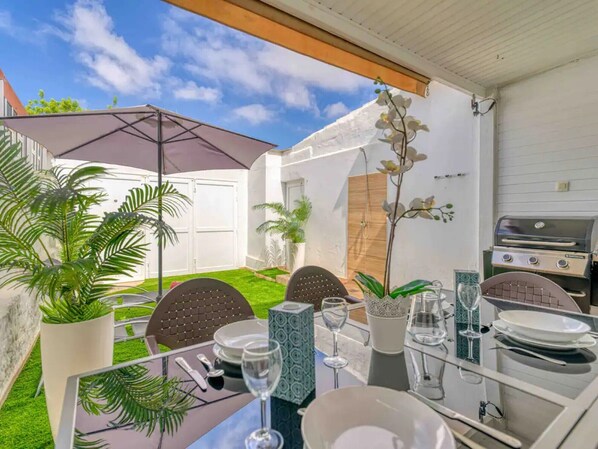 An outdoor dining area with a table set for four, surrounded by chairs, a grill, and potted plants.