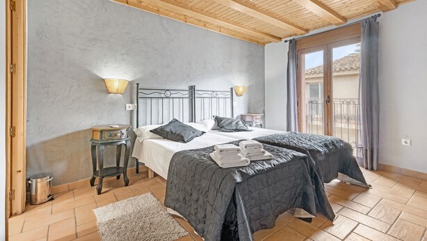 A bedroom with a wooden ceiling, two beds with dark bedding, bedside lamps, a small table, and a view of the outside through a window.