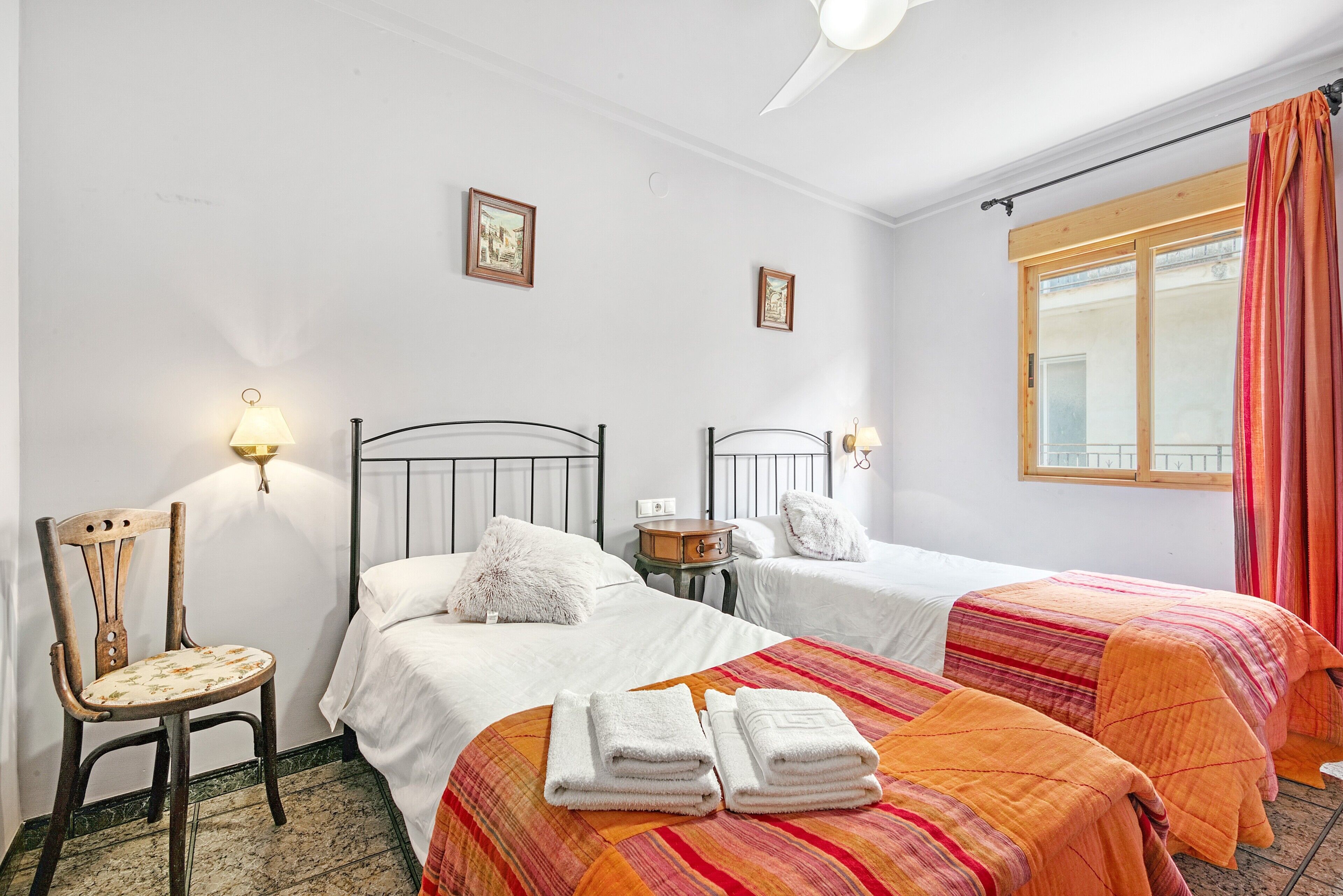 Hotellihuone, jossa on kaksi sänkyä, tuoli, lamppu ja ikkuna, jossa on verhot.