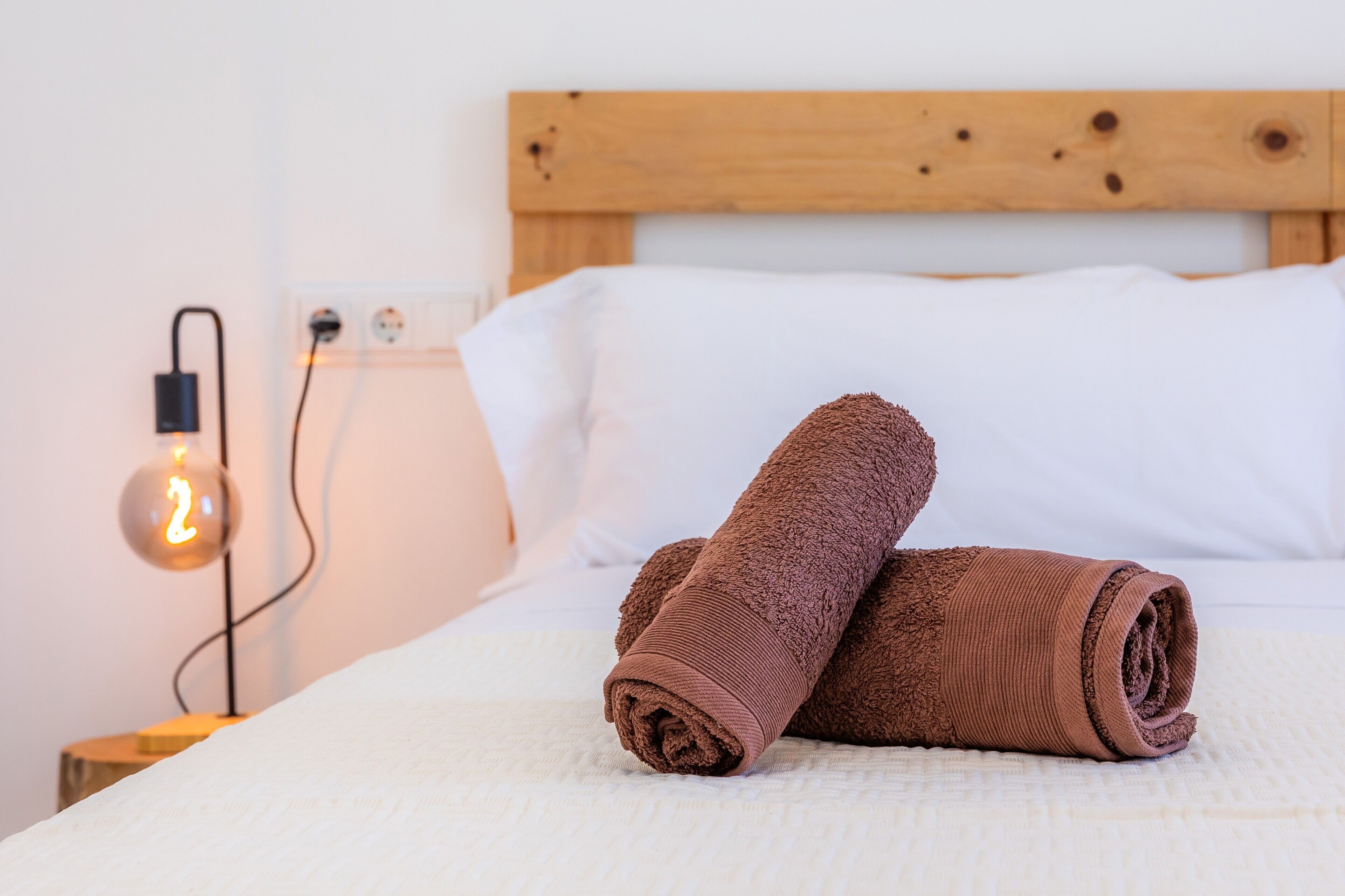 Un letto ben rifatto con un poggiatesta in legno, una lampada da comodino e un asciugamano arrotolato sul letto.