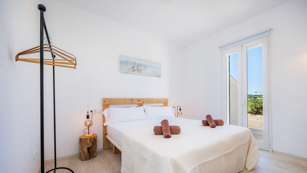 Un dormitorio con cama, cabecera de madera, un cuadro, una lámpara de pie y vistas por una puerta.