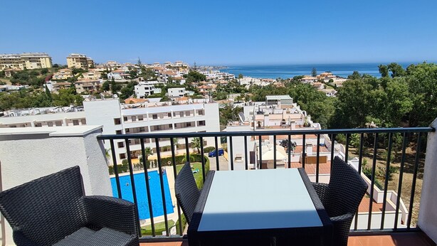 A balcony with wicker furniture, a pool, and a view of the sea and town. - Apartment 'Casa Pals La Cala' with Shared Pool, Wi-Fi and Air Conditioning (La Cala de Mijas)