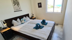 A neatly made bed with patterned bedding, a bedside lamp, a small table with a plant, and a window with a view. - Apartment 'Casa Pals La Cala' with Shared Pool, Wi-Fi and Air Conditioning (La Cala de Mijas)