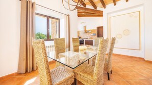 A dining area with a glass table and wicker chairs, a kitchen in the background, and a view through a window. - Holiday Home 'Na Berganta' with Private Terrace, Private Garden and Wi-Fi (Illes Balears)