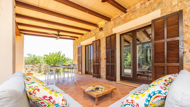 An outdoor patio with a wooden table, two sofas with colorful pillows, and a stone wall. - Holiday Home 'Na Berganta' with Private Terrace, Private Garden and Wi-Fi (Illes Balears)