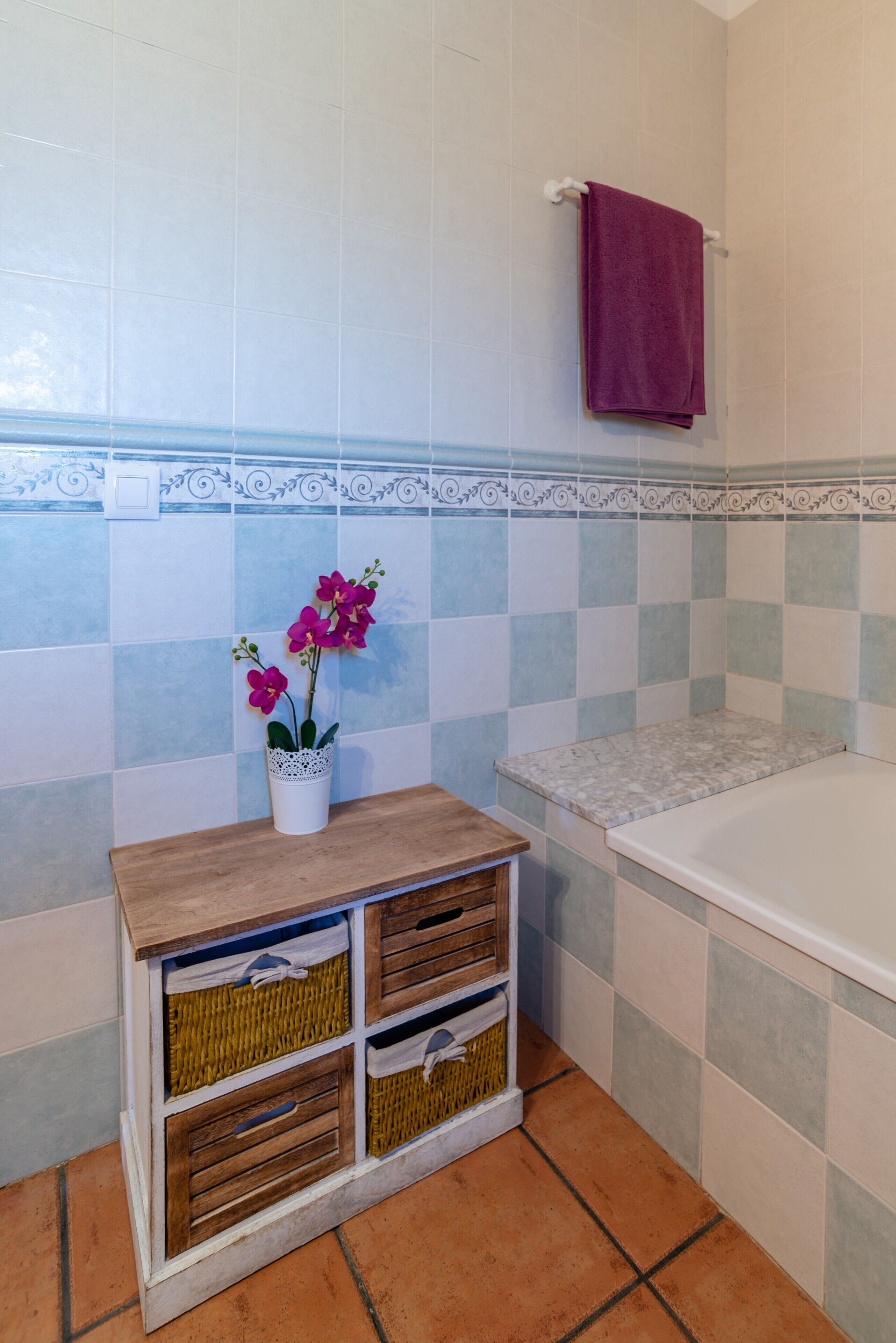 Une salle de bain avec un mur carrelé, un meuble-lavabo en bois doté de paniers, une serviette violette et une baignoire.