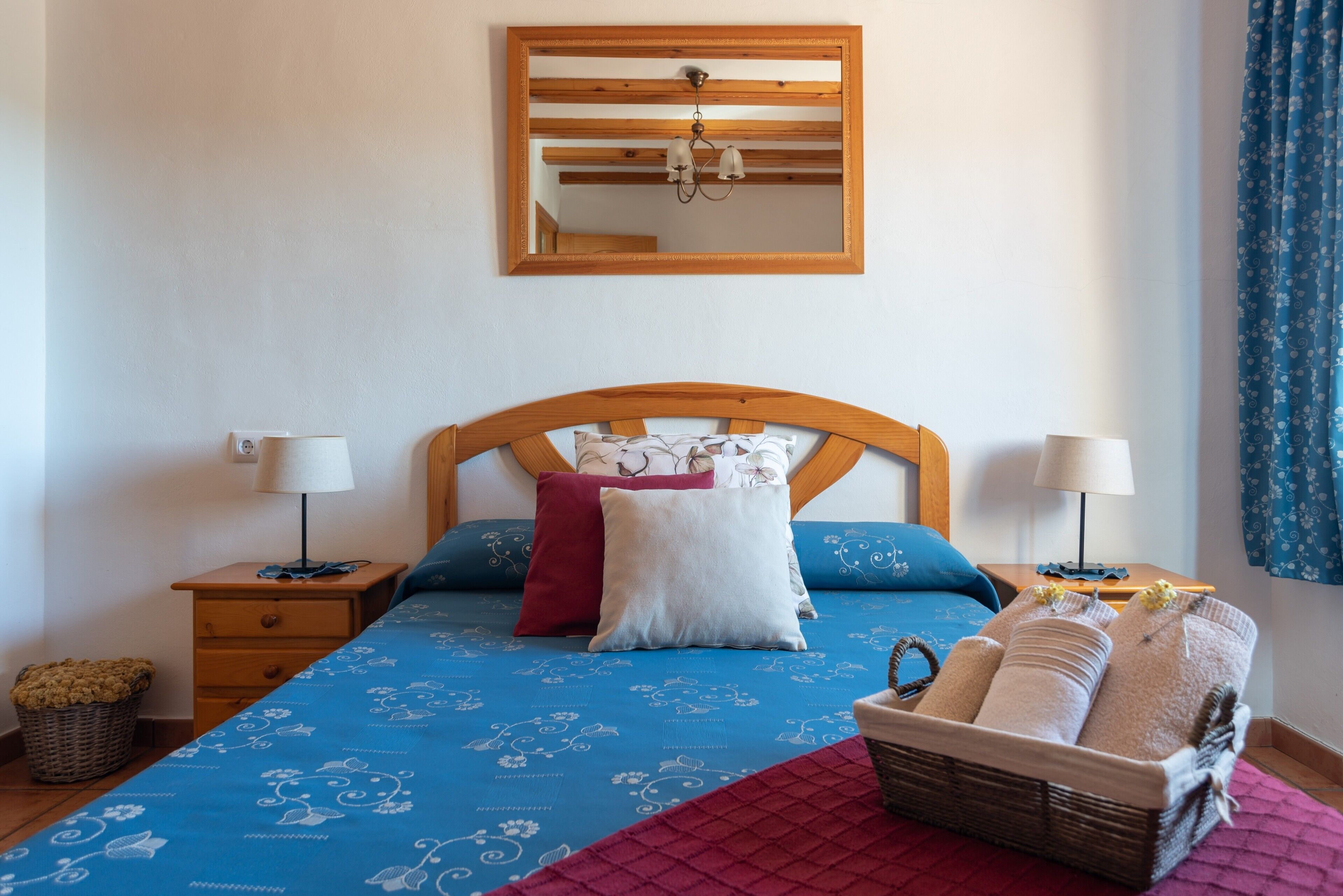 A bedroom with a wooden headboard, a bed with patterned bedding, two bedside lamps, a mirror, and a basket of towels.