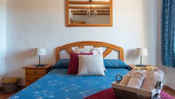 A bedroom with a wooden headboard, a bed with patterned bedding, two bedside lamps, a mirror, and a basket of towels.