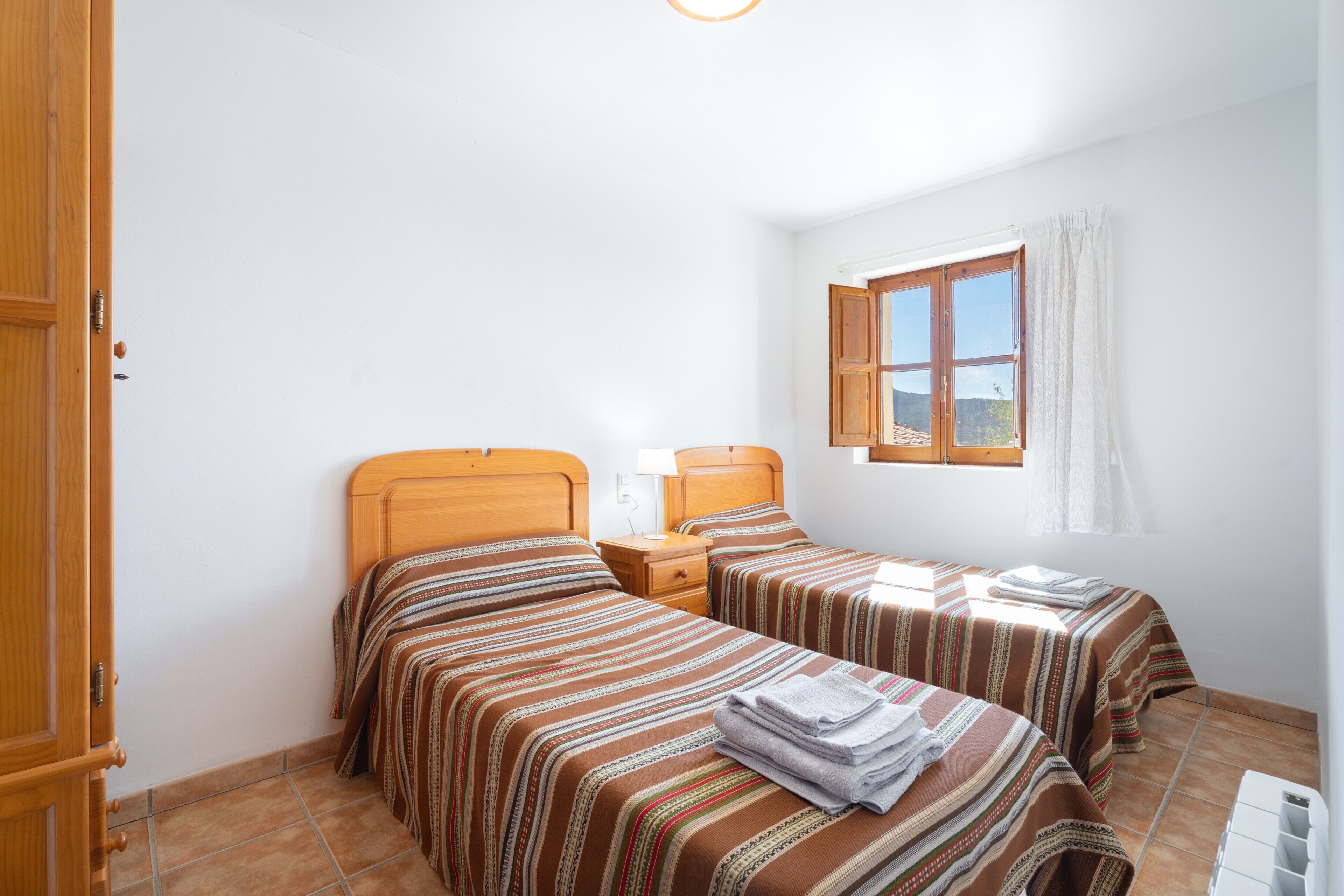 A bedroom with two beds, a window with curtains, a wooden door, and a tiled floor.