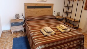 A bedroom with a wooden headboard, a bed with striped bedding, a nightstand with a lamp, a small shelf, and a wardrobe with hangers. - Apartment 'Antiga Escola De Molinell' with Mountain View, Private Terrace and Private Garden (Culla)