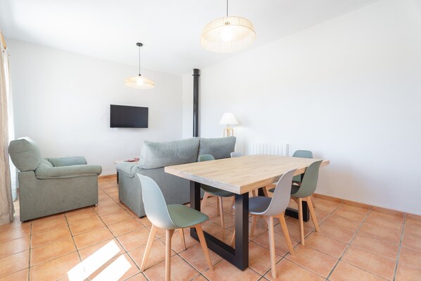 A modern living room with a wooden dining table, grey sofas, and a flat-screen TV mounted on the wall.