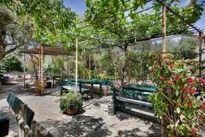 A covered outdoor seating area with stone flooring, green benches, and potted plants.